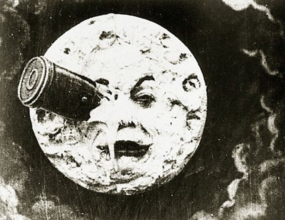A round, cratered Moon with a human face grimaces in a dark, cloudy sky, a bullet-shaped capsule lodged in one of its eyes.