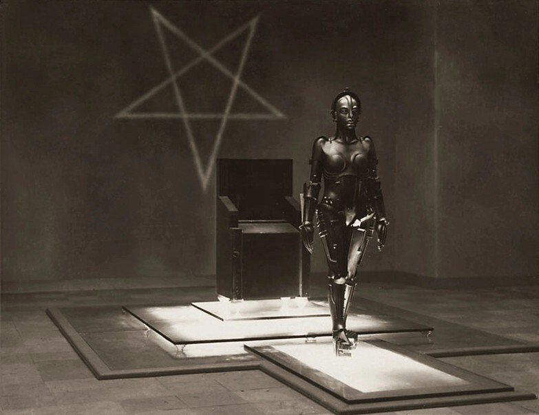 A metallic humanoid robot stands with one knee raised on a lit platform in a bare, dim hall, in front of a dark chair, with a five-pointed star drawn on the wall behind it, in a sepia photograph.