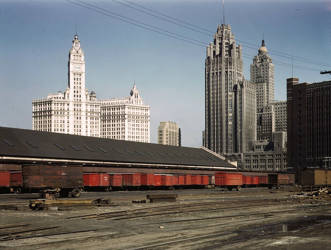 Red freight cars and a low freight house in a rail yard, with the white Wrigley Building and the Gothic Tribune Tower rising behind.