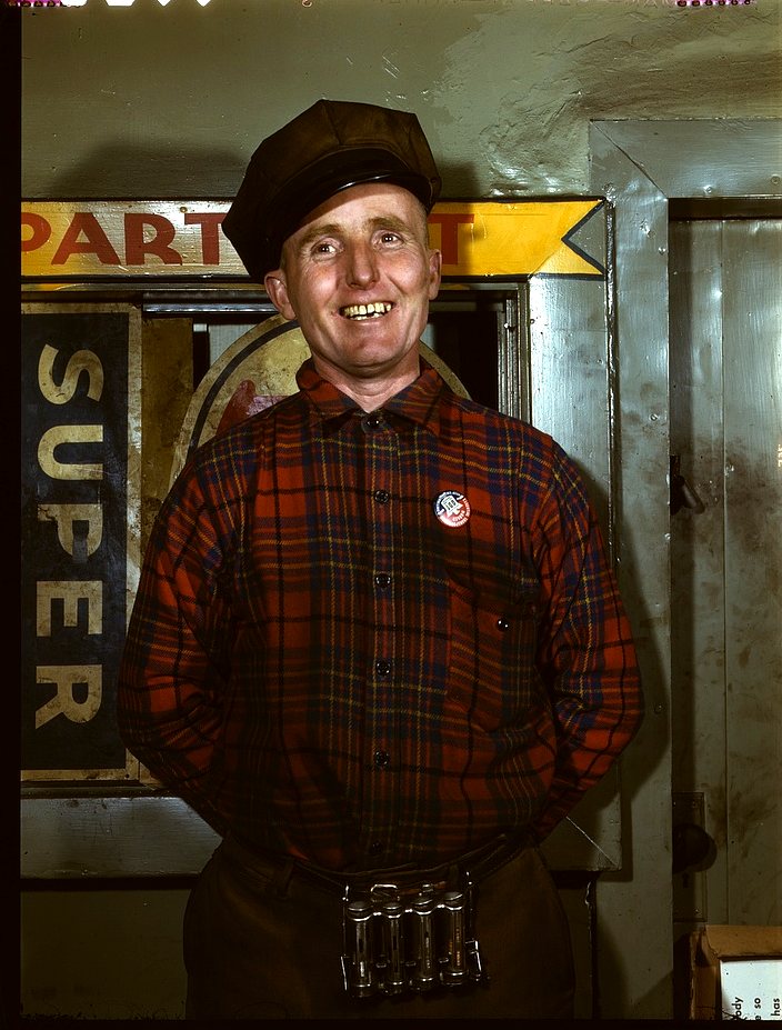 A smiling man in a plaid shirt and cap stands in a garage doorway with a small badge on his chest and tools at his belt.