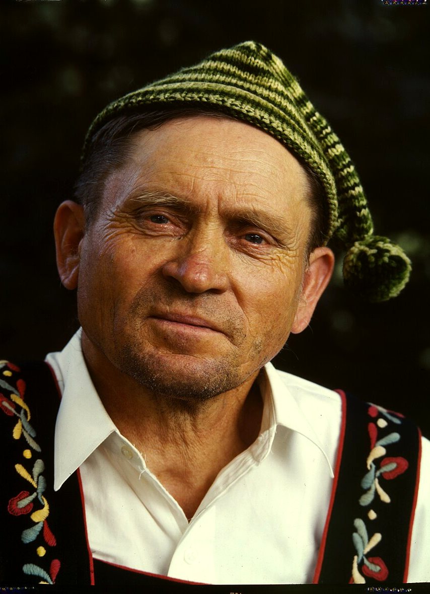 Close portrait of a man in a green knitted cap with a pompom, a white collared shirt and an embroidered black vest.