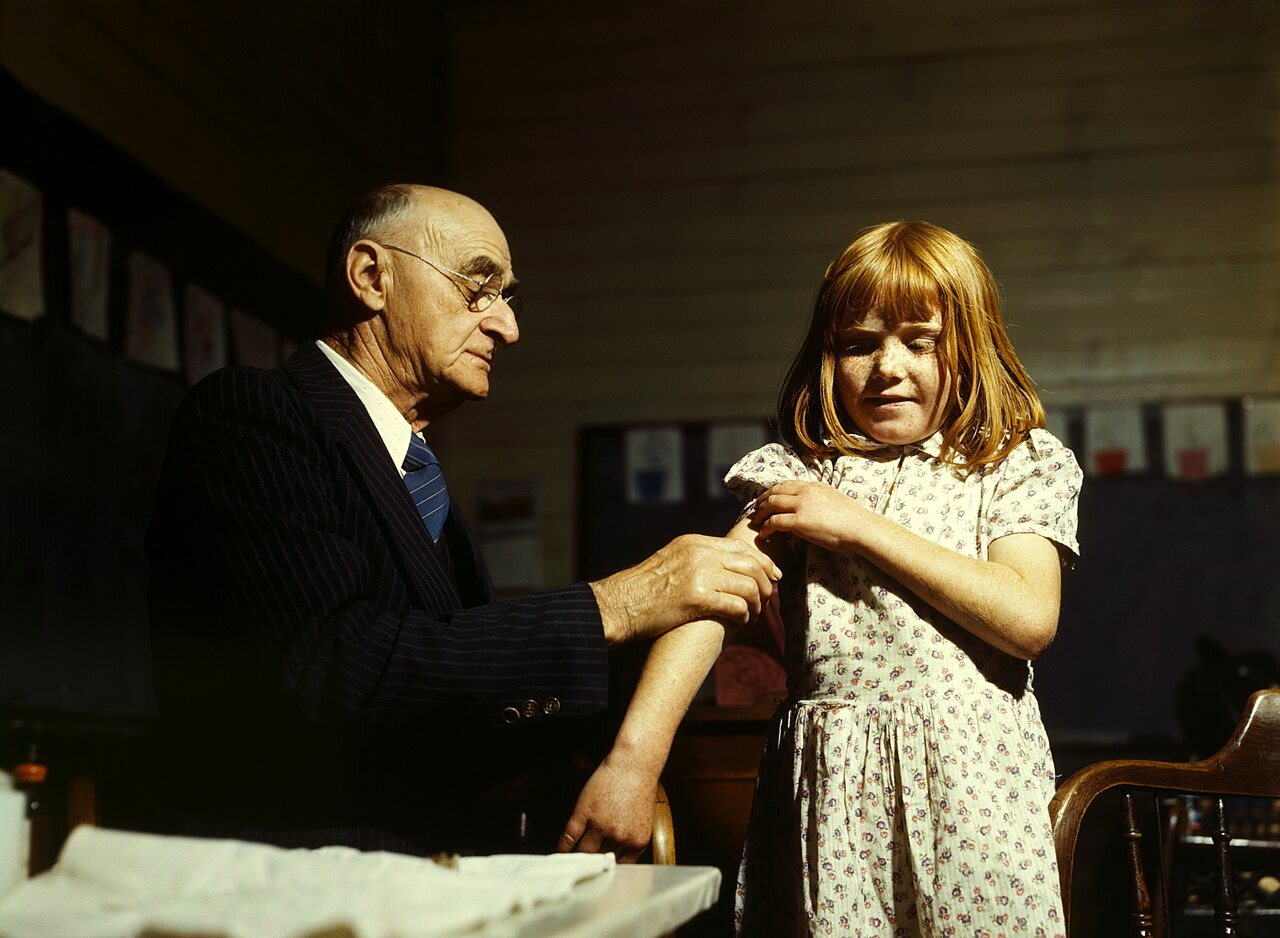 An older doctor in a dark suit gives a young girl an injection in the upper arm while she squints and holds up her sleeve.
