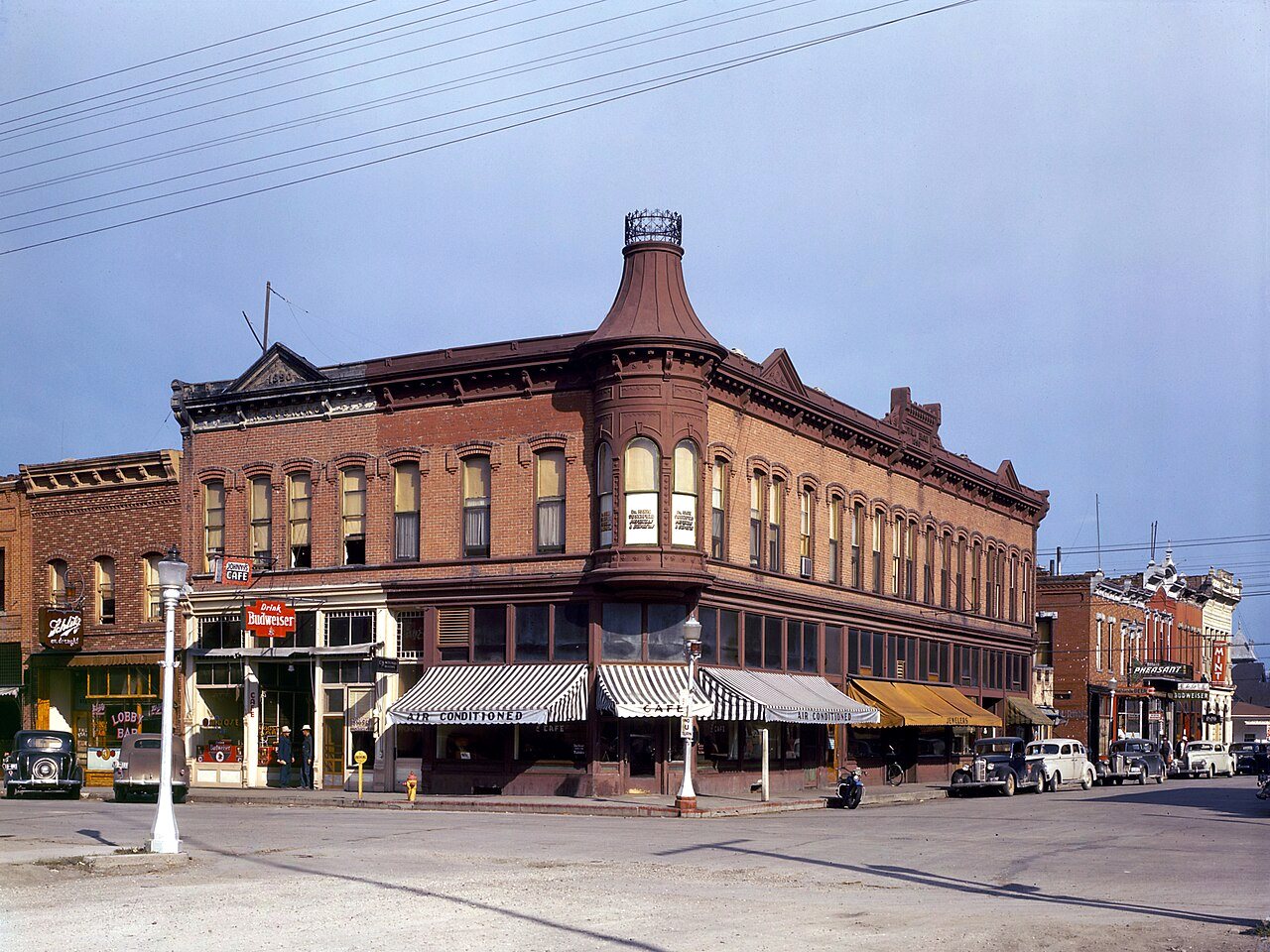 A brick corner building with a turret and striped awnings on a wide, nearly empty street in a small Montana town.