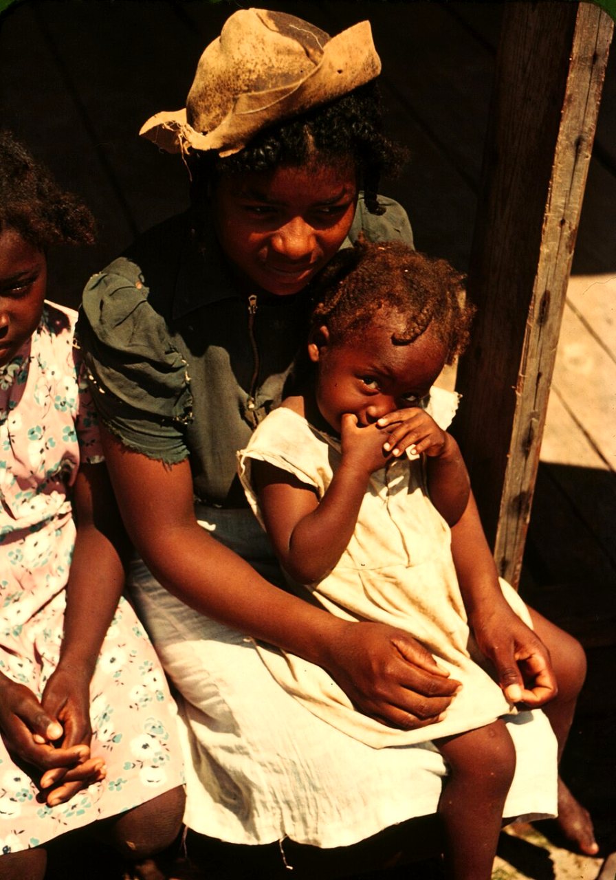 A girl in a straw hat holds a smaller child in a pale dress on her lap on a shaded porch, with another girl in a floral dress beside them.