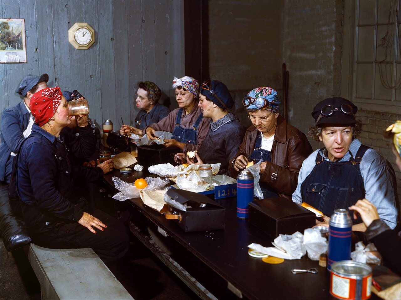 Women in coveralls and head scarves eat lunch around a long table in a railroad roundhouse rest room.