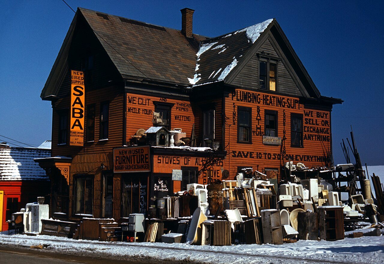 An orange-brown building with signs for plumbing, heating and furniture, with used stoves and fixtures stacked in front under a deep blue sky.
