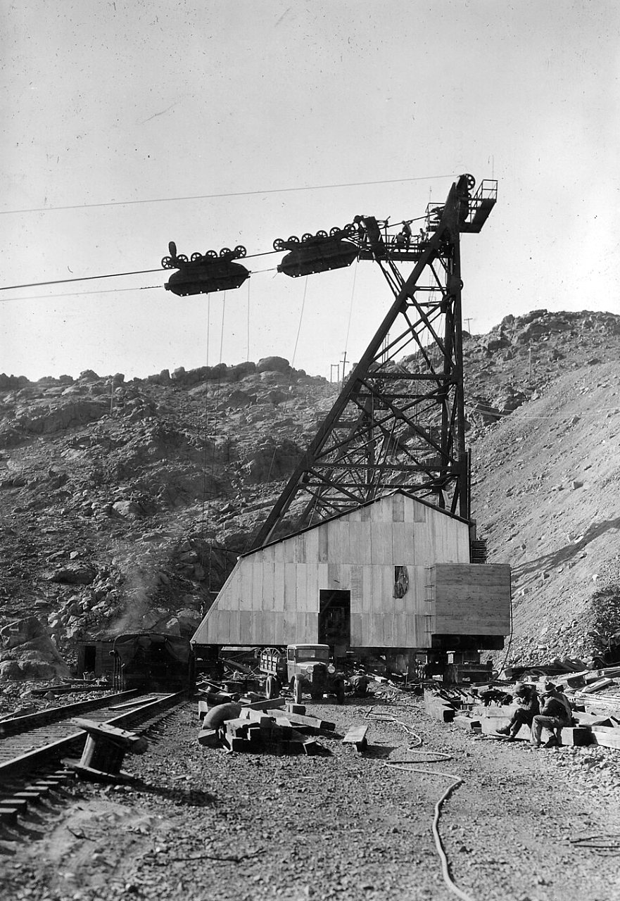 A tall steel cableway tower stands beside a shed in a rocky canyon, with two loads hanging from the cables overhead.