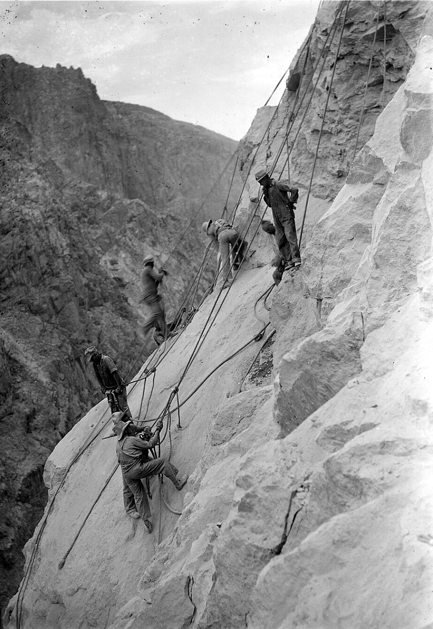 Men hang from ropes on a steep, pale rock wall of a canyon high above the river.