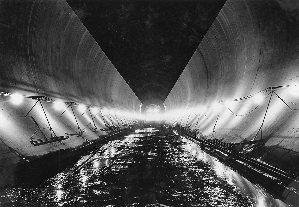 A long concrete-lined tunnel with lights along its curved walls and a wet floor reflecting them.