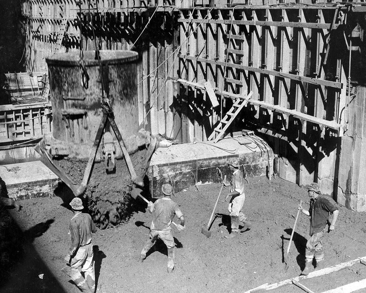 Workers in hard hats stand with long-handled tools on fresh concrete beside a wooden form as a bucket empties.