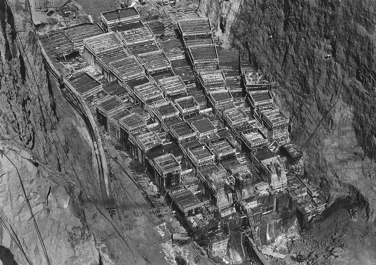 A telephoto view from above of the dam under construction, a grid of separate concrete blocks and wooden forms in the canyon.