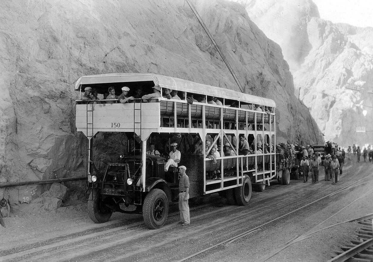 A two-level open-sided coach packed with men drives through a rock cut, with other workers walking beside it.
