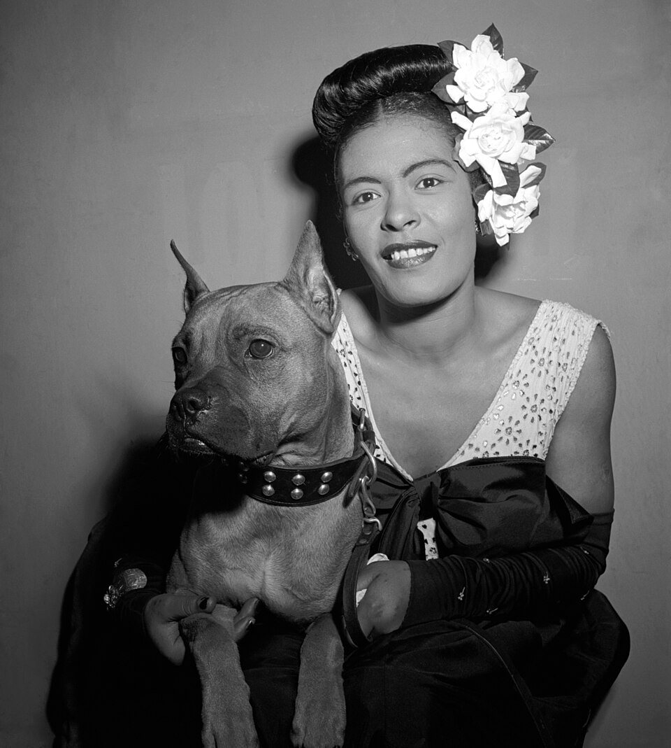 Singer Billie Holiday, with white flowers in her hair, sits with her boxer dog Mister, who wears a studded collar, in front of a plain backdrop.