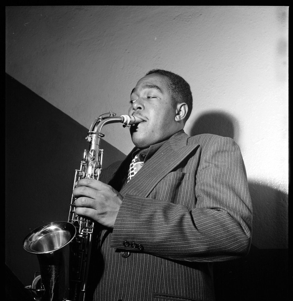 Charlie Parker plays an alto saxophone in a pinstripe suit, with a bright wall and his shadow behind him.