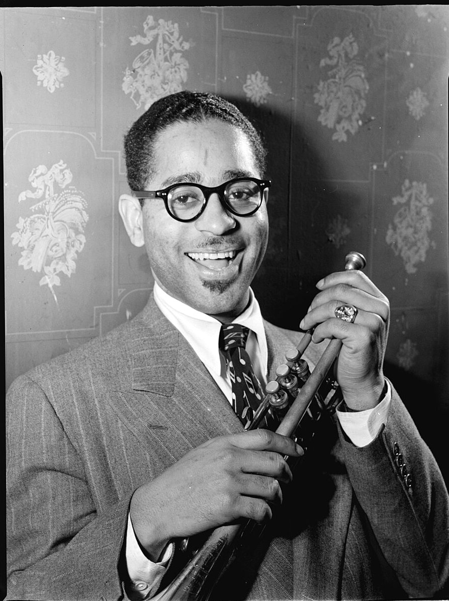 Dizzy Gillespie in round black glasses and a pinstripe suit smiles while holding a trumpet in front of a flowered wall.