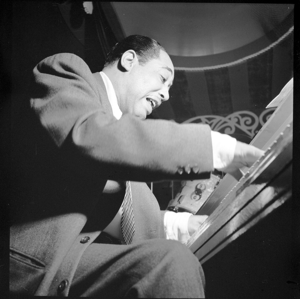Duke Ellington leans over a piano keyboard with his mouth open, caught mid-phrase, with a striped awning behind him.