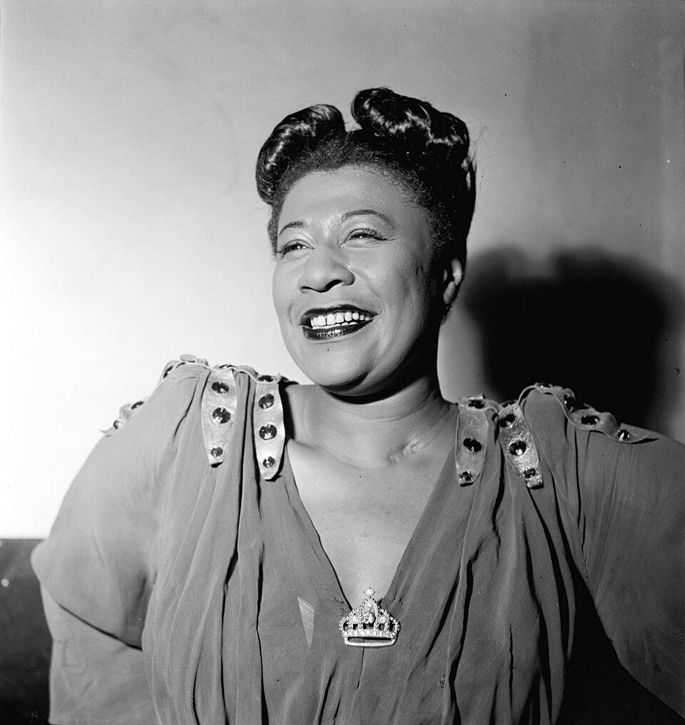 Ella Fitzgerald laughs with her head turned to one side, wearing a dress with studded shoulders and a small crown pin.