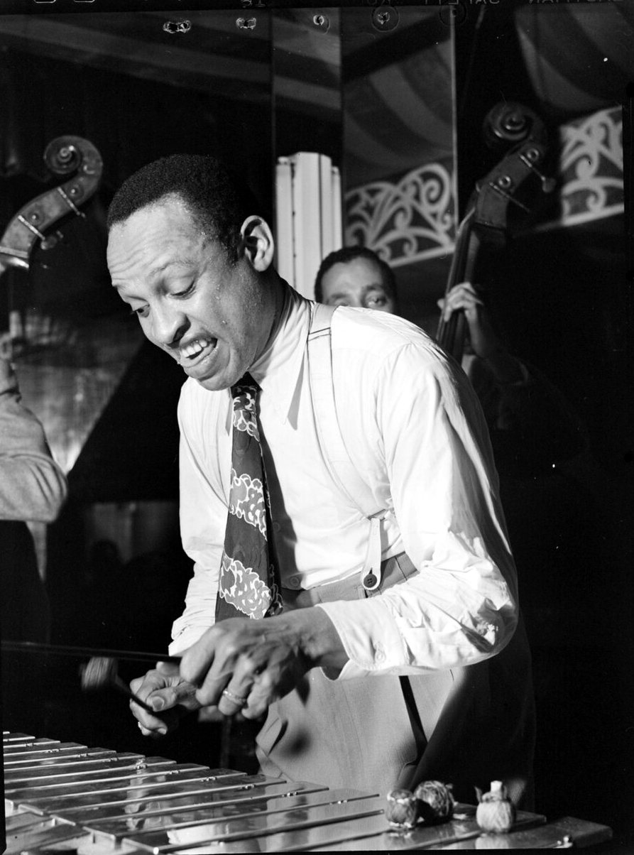 Lionel Hampton, in a white shirt and patterned tie, leans over a vibraphone with his mouth open as a double bass shows behind him.