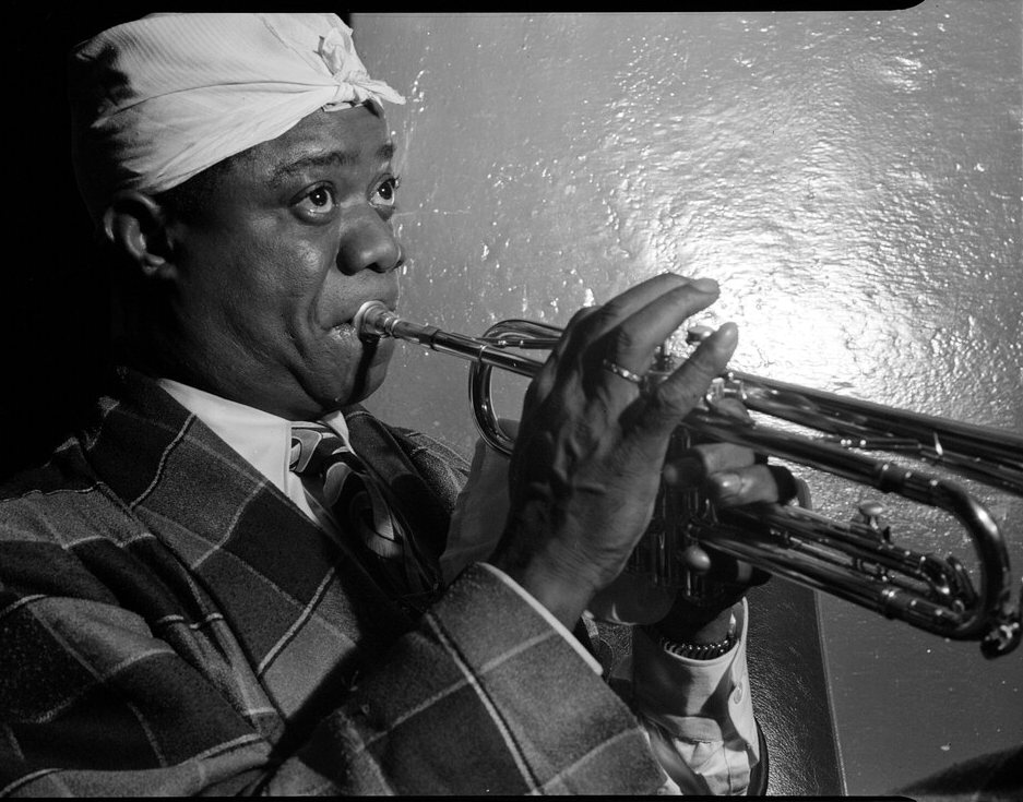 Louis Armstrong, in a plaid jacket and a white cloth on his head, plays the trumpet in front of a reflective wall.
