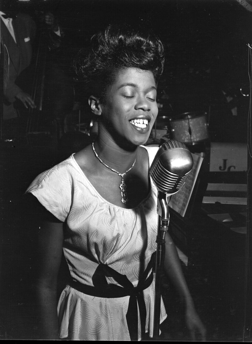 Sarah Vaughan sings with her eyes closed beside a vintage microphone, wearing a light dress with a dark ribbon at the waist.