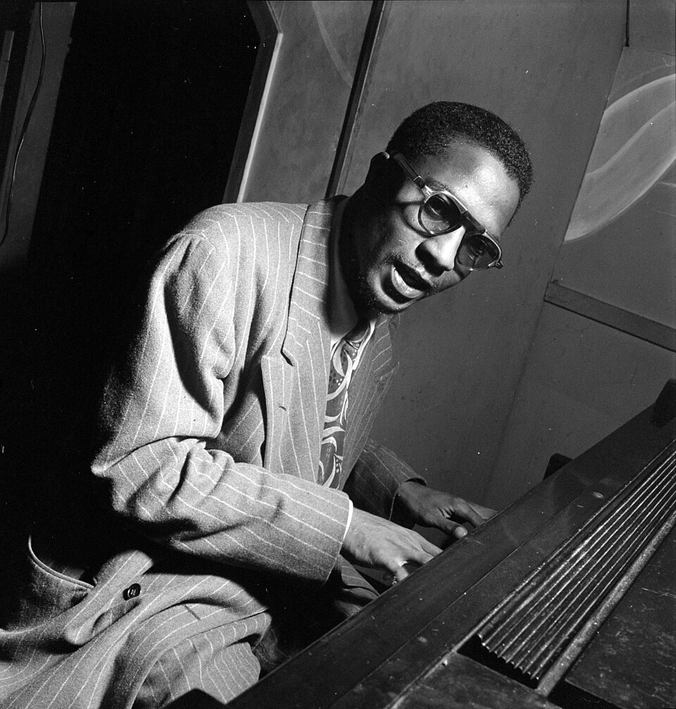 Thelonious Monk in glasses and a striped jacket sits sideways at an upright piano, looking toward the camera.