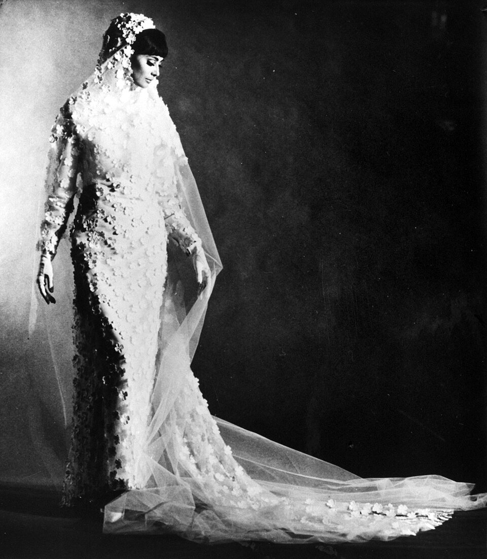 A model in a long bridal gown covered in small flowers, with a veil falling to a long train, stands against a dark background.