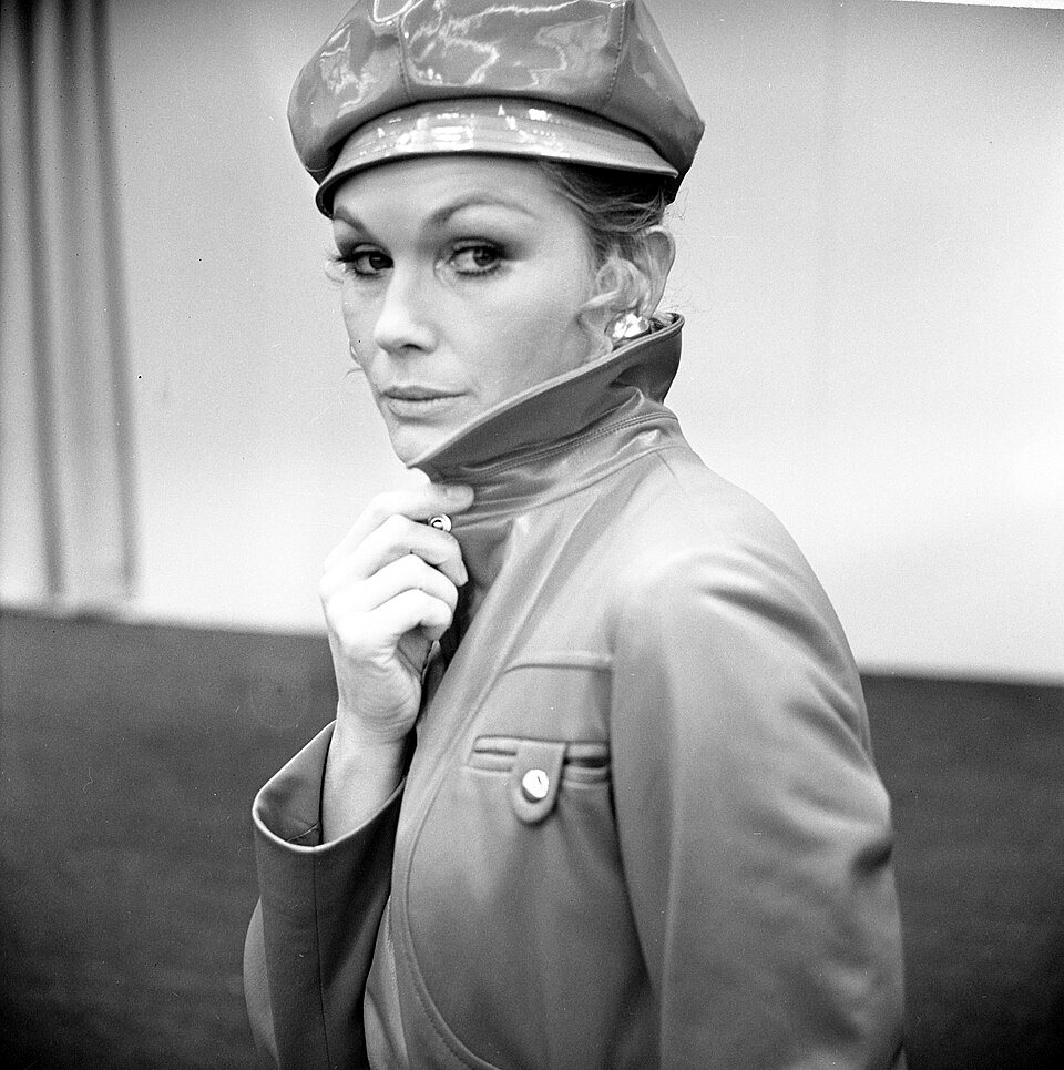 A woman in a glossy patent-leather cap and a matching high-collared coat looks sideways at the camera with one hand at her collar.