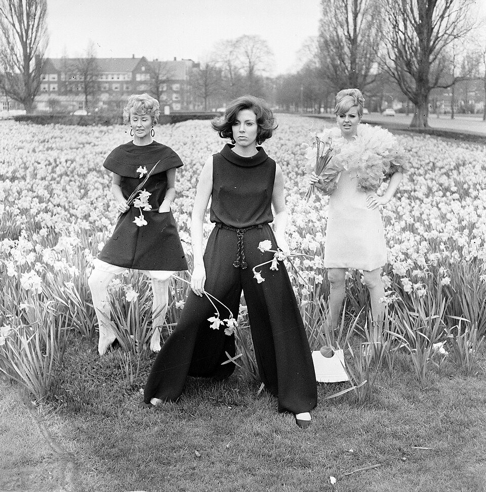 Three women in autumn outfits stand in a bed of daffodils in a park with bare trees behind them: a dark short tunic, a sleeveless top with very wide trousers, and a pale feathered mini dress.