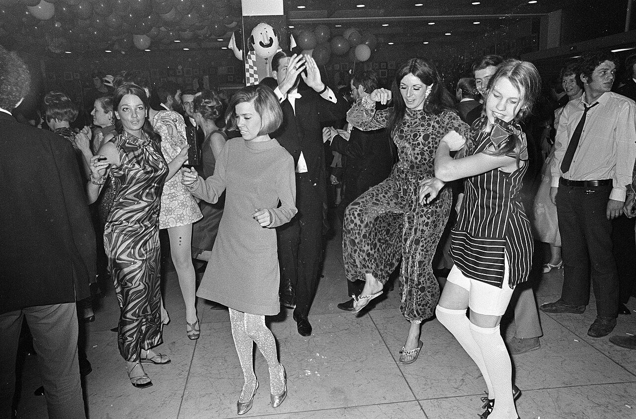 Women in short patterned and striped dresses, patterned tights and white knee socks dance in a dim, crowded hall, among men in suits.