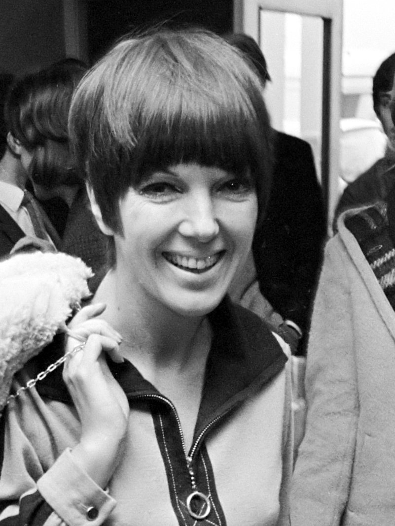 A smiling woman with a sharp dark bob and a straight fringe, wearing a zip-front jacket with a ring pull, holds a fluffy white item at her shoulder in a crowd.
