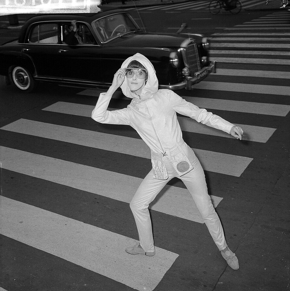 A model in a pale hooded jacket, trousers and tinted glasses poses on a zebra crossing, one hand on her hood and the other arm flung out, with a dark car driving past behind her.