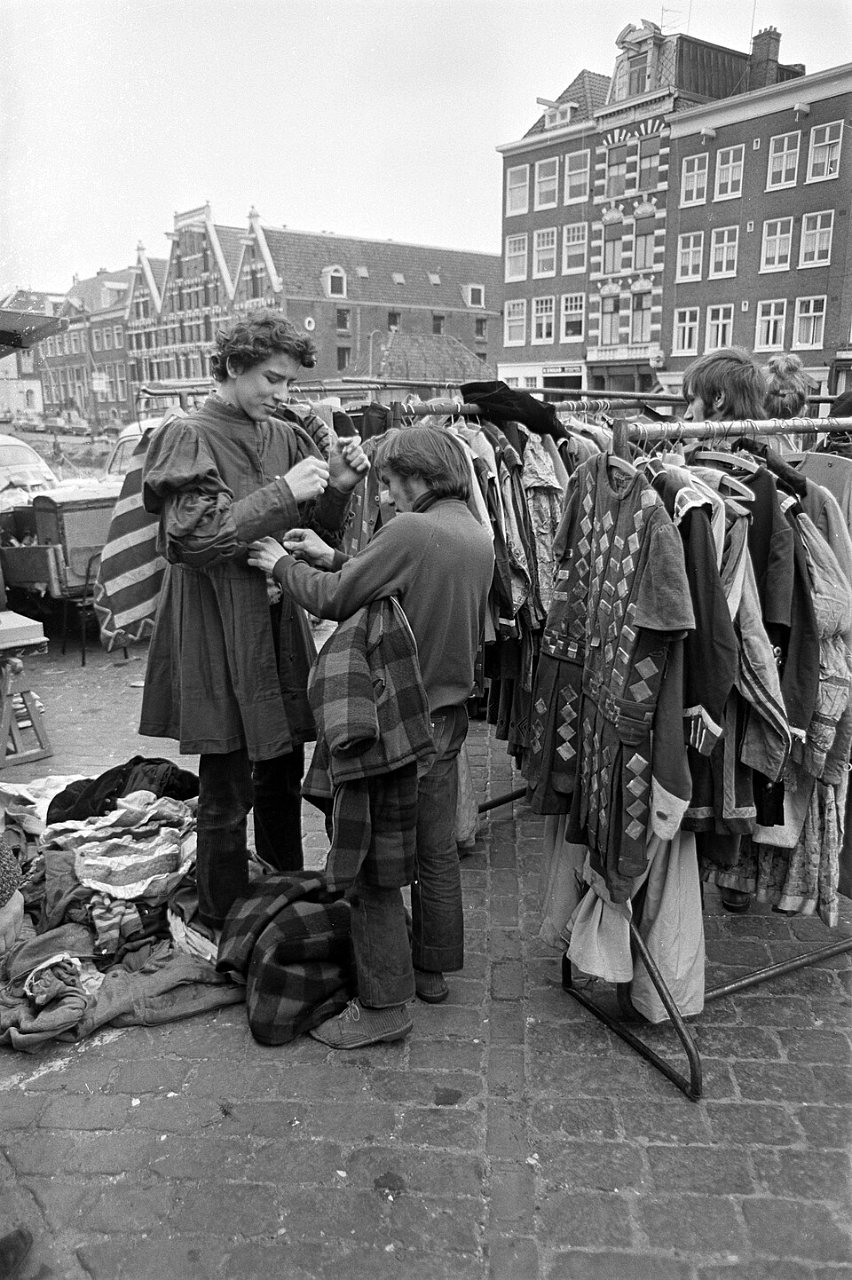 A man in a long dark tunic and a shopper in a plaid coat stand at a crowded clothes rack on a cobbled square, with Amsterdam canal houses behind them.