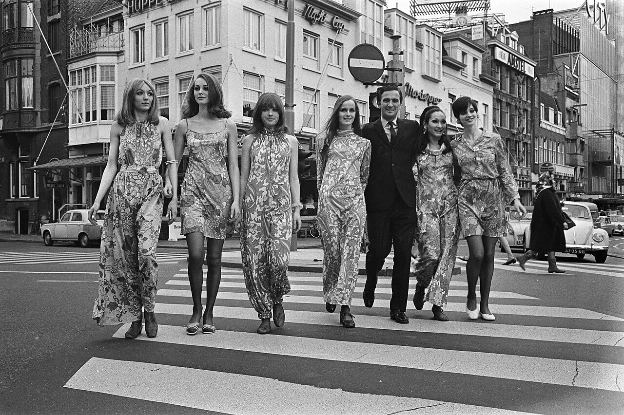 Six women in matching leaf-and-flower print dresses of different lengths walk across a zebra crossing beside a man in a dark suit, with tall brick buildings behind them.