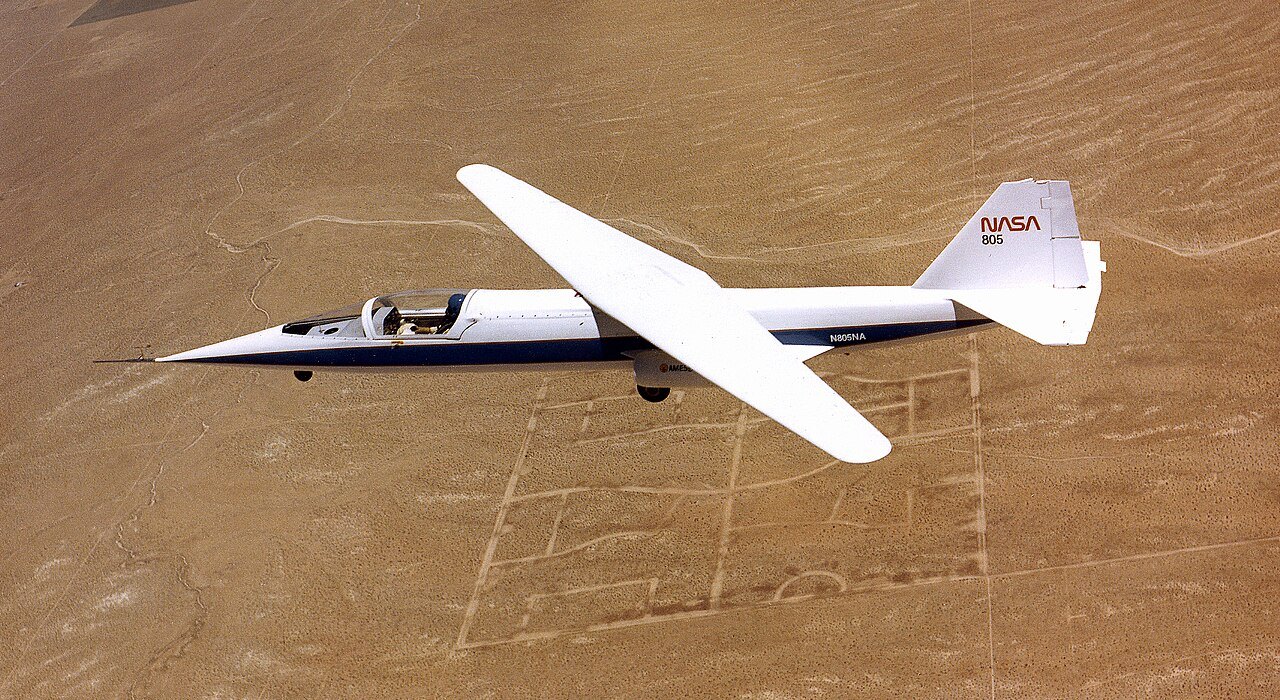 A small white research jet flies over brown desert with its single long wing turned at an angle, one tip swept forward and the other back.