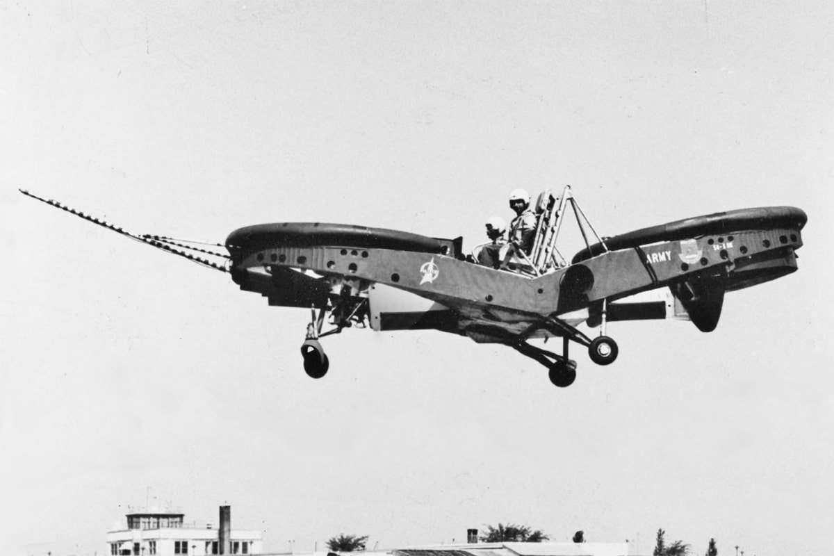 A flat, boxy aircraft with a ducted rotor at each end hovers low over the ground with two crewmen aboard, a thin line trailing from its nose to the left.
