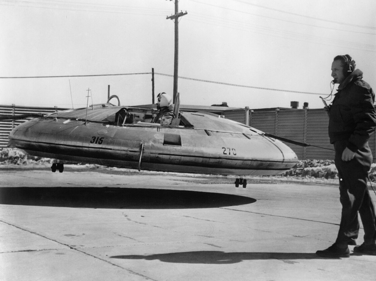 A round, saucer-shaped aircraft hovers just above the ground while a man in a dark jacket stands beside it, with a power pole and a low building behind them.