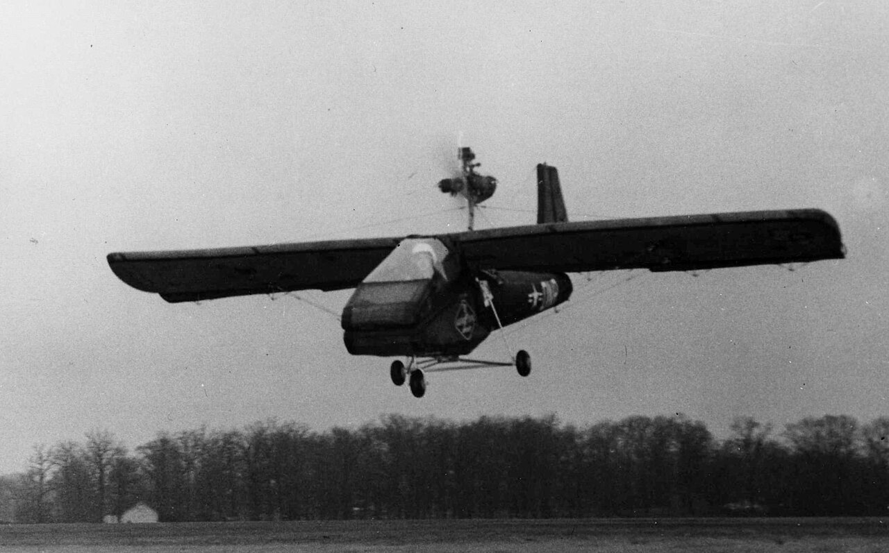 A small high-winged aircraft with a pod-shaped cockpit and a tail on a boom comes in low over the ground, with a line of trees behind it.