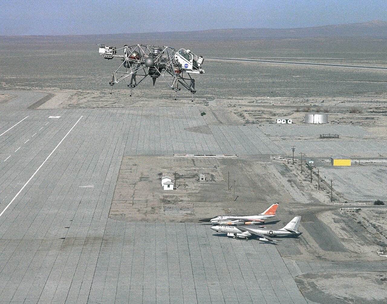 A spindly four-legged open-frame vehicle hovers high above a flat desert airfield, with parked aircraft small on the ground below.