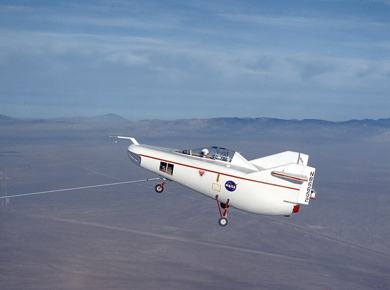 A small white wingless aircraft with a rounded body and twin tail fins flies low over a desert lakebed, pulled along on a long tow line.