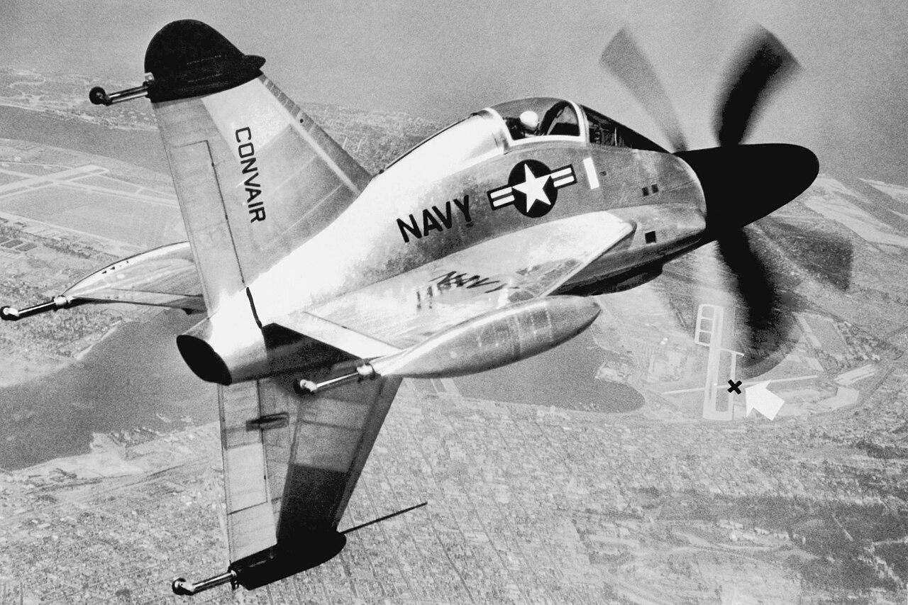 A Navy tail-sitter fighter marked NAVY banks steeply over a dry landscape, its propeller blurred at the nose and its wings seen from below.