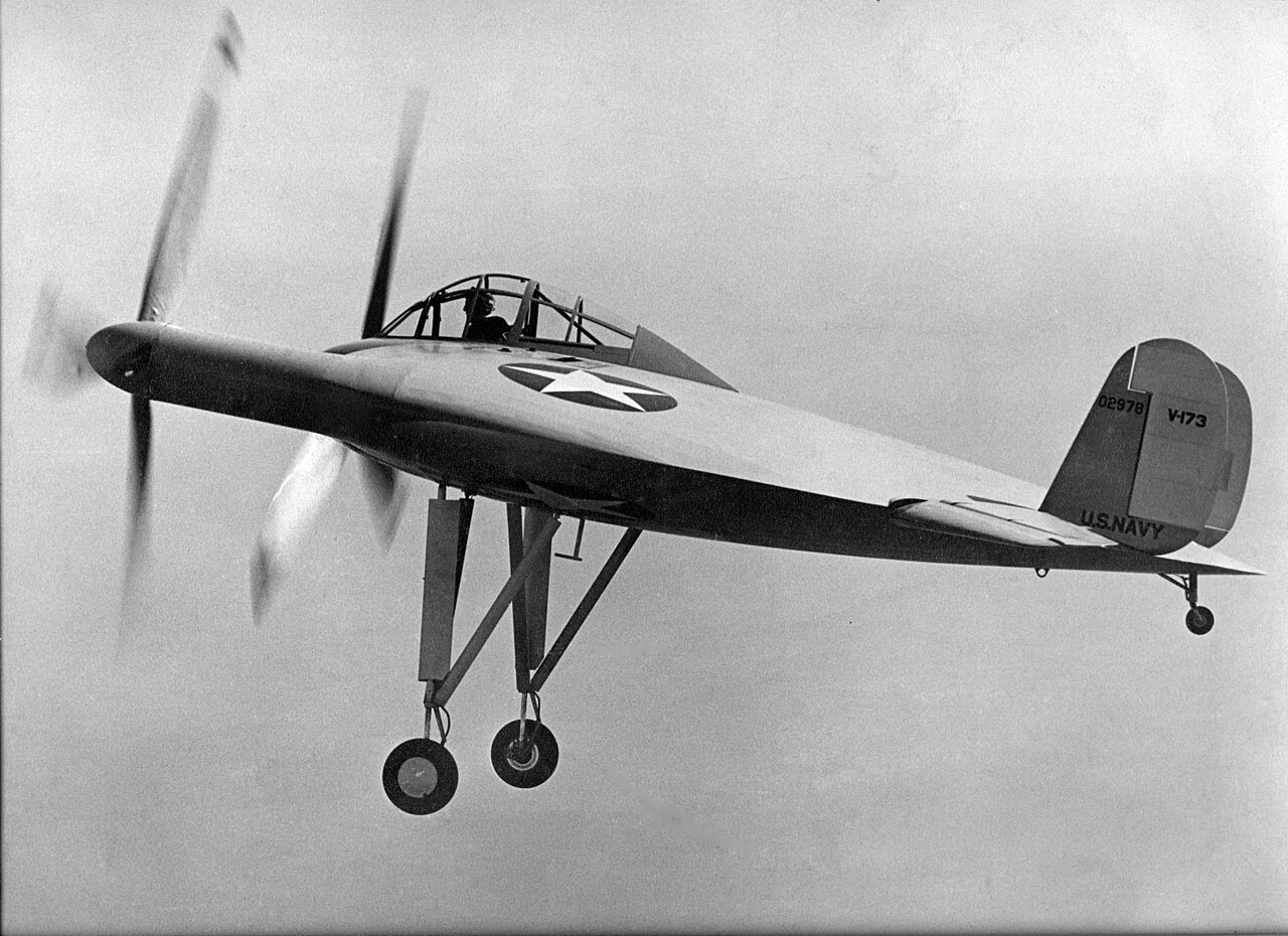 A disc-shaped experimental aircraft banks in flight, its two large propellers standing out at the front edge of the wing and a small tail fin at the right, against a pale sky.