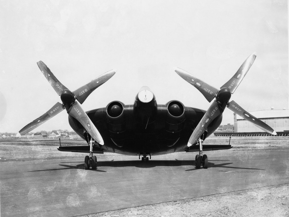 A stubby round-winged aircraft sits on a paved runway seen head-on, with two huge propellers angled outward above its wing.