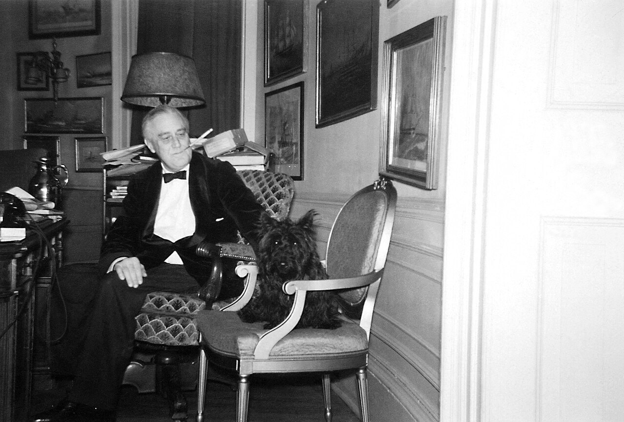 An older man in a dark suit sits in an armchair in a study beside a small black Scottish terrier, with a lamp and framed pictures on the wall behind them.