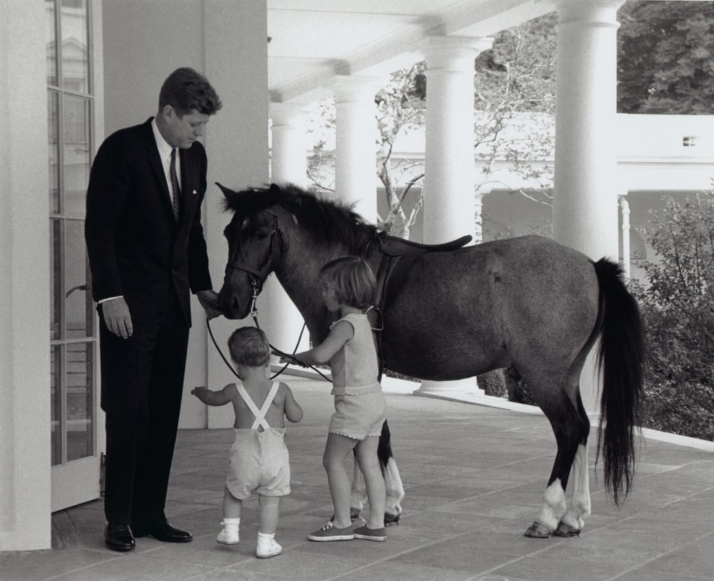 A man in a dark suit stands beside a brown pony on a white-columned walkway while two small children in light clothes stand in front of it.