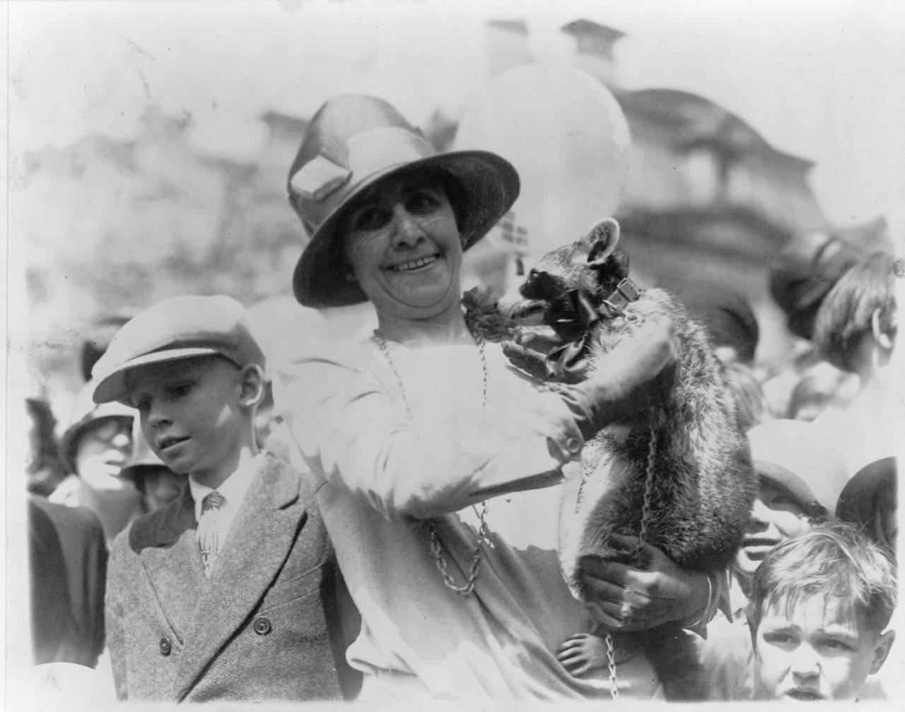 A woman in a wide-brimmed hat holds a raccoon up beside her shoulder while a boy in a cap and other children crowd close on a lawn.