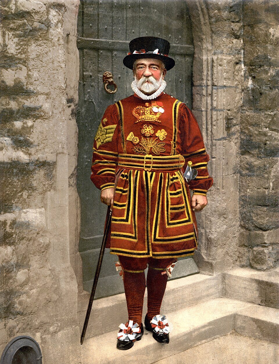 A white-bearded Yeoman Warder in a red and gold Tudor state uniform and black hat stands on stone steps in front of a green door, holding a staff.