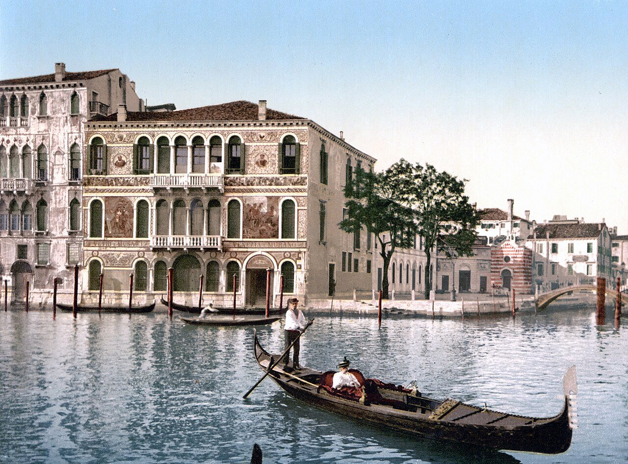 A decorated palace with arched windows stands on the Grand Canal in Venice as a gondolier poles a gondola with a passenger past it.
