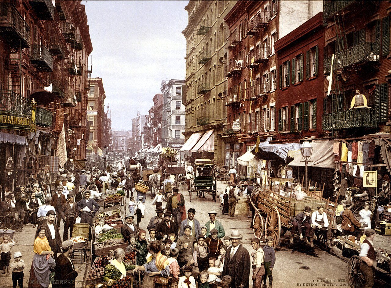 A crowded street lined with tenement buildings, pushcarts and market stalls, with people filling the roadway.