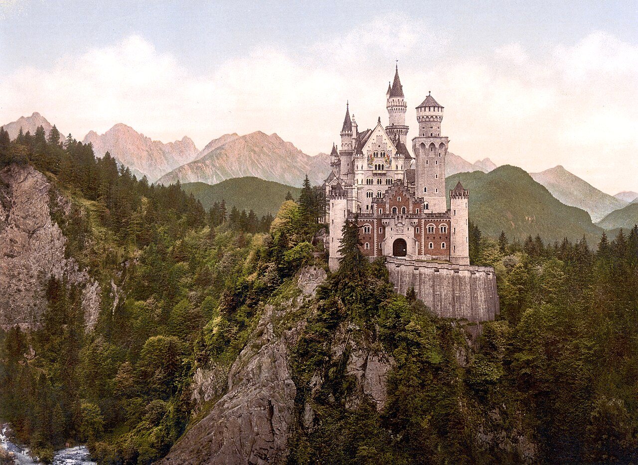 A white castle with many towers sits on a rocky ridge above dark forest, with mountains and a pale sky behind.