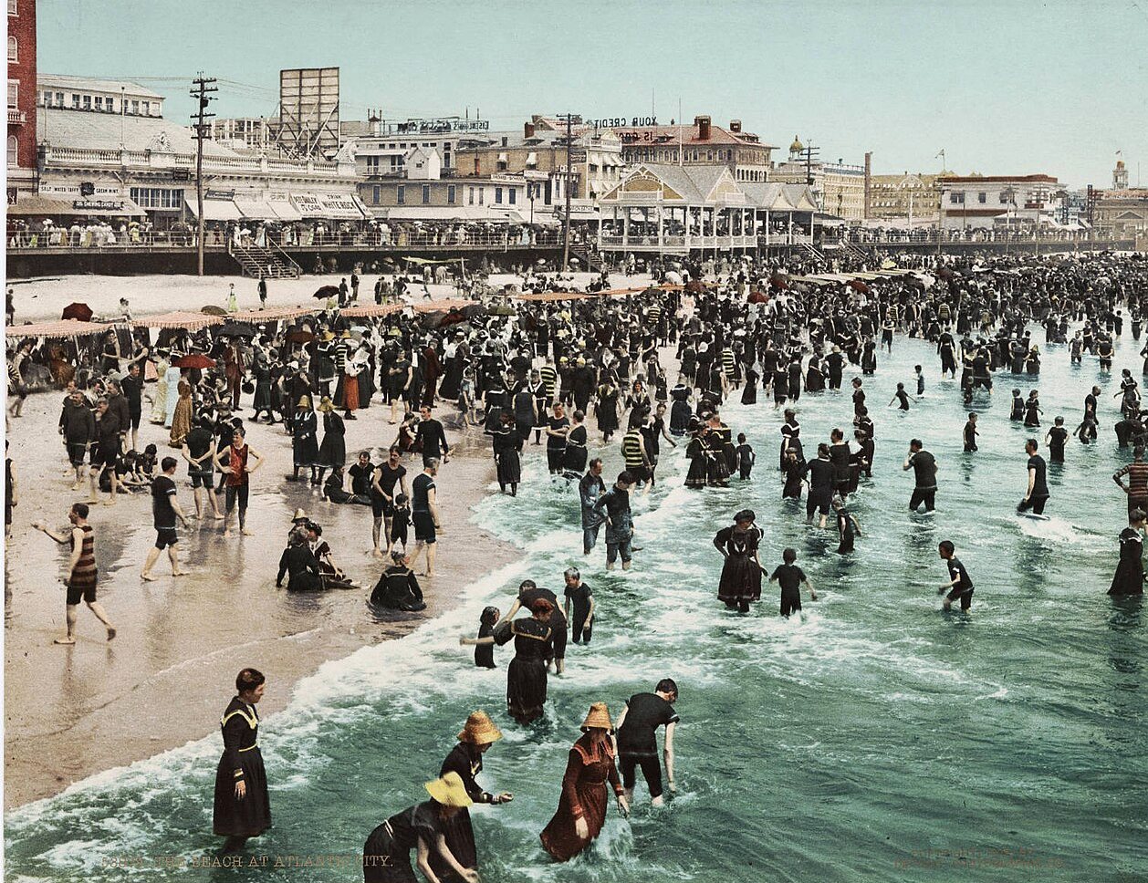 Hundreds of bathers in dark bathing costumes wade and stand in the surf at a crowded beach, with a boardwalk and hotels behind.